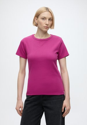 Kvinde med kort blondt hår iført en tætsiddende magenta T-shirt med lille "HUGO" logo og sorte bukser, stående mod en ensfarvet hvid baggrund.