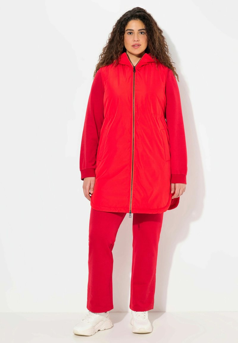 Rote gepolsterte Jacke mit einem Reißverschluss auf der Vorderseite, Kapuze und langen Ärmeln, kombiniert mit passenden roten Hosen und weißen Turnschuhen. Glatte Textur insgesamt.