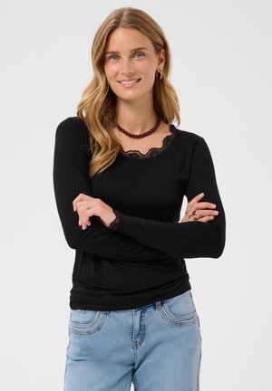 Frau mit hellbraunen Haaren, die ein schwarzes Langarmoberteil mit Spitzenbesatz und hellblaue Jeans trägt, lächelnd mit verschränkten Armen vor weißem Hintergrund.