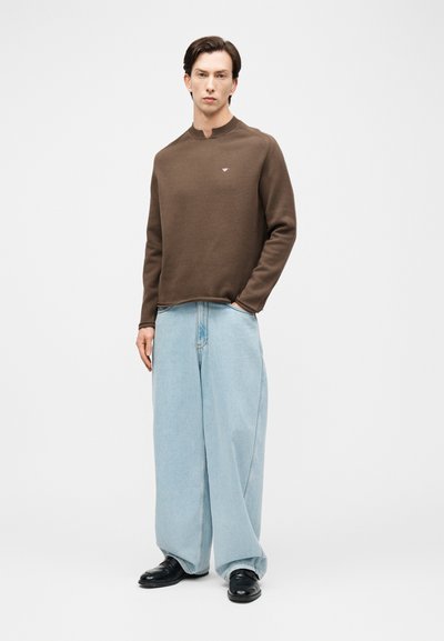 Άνδρας που φοράει καφέ πουλόβερ με μακριά μανίκια, ανοιχτό μπλε τζιν παντελόνι καμπάνα και μαύρα παπούτσια, στέκεται με το ένα χέρι στην τσέπη, σε λευκό φόντο.