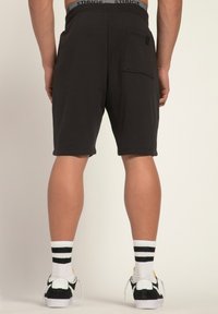 Schwarze Shorts mit entspannter Passform, ausgestattet mit einer Gesäßtasche, weichem Stoff und einem elastischen Bund mit 'STHUGE'-Logo. Getragen mit schwarz-weißen Schuhen.