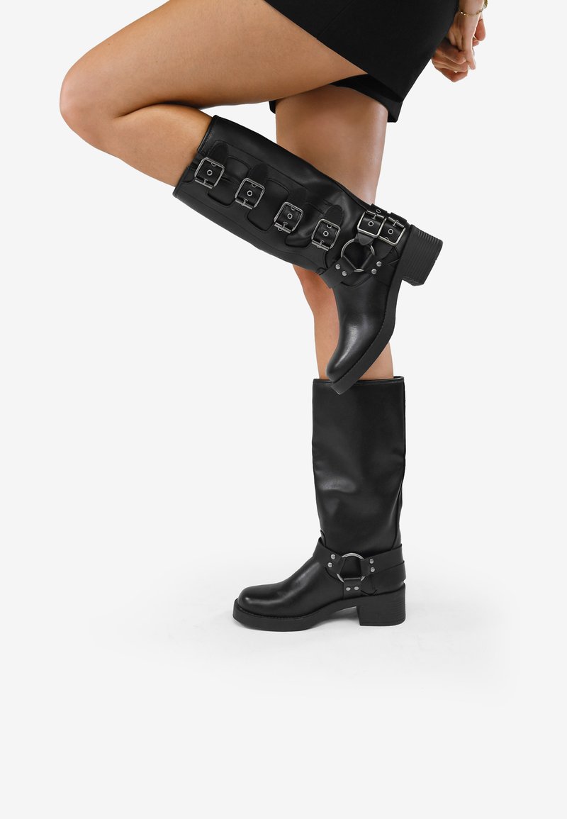 Botas negras hasta la rodilla hechas de material suave, con múltiples hebillas plateadas y un tacón grueso. Una bota está doblada hacia abajo.