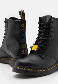 Botas de combate negras con una textura estampada, acentos amarillos y gráficos multicolores en el lateral. Presentan un sistema de bloqueo de cordones amarillo y costuras en contraste.