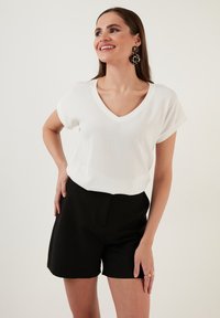 Top bianco a V con texture e maniche corte arrotolate, abbinato a pantaloni corti neri a vita alta. Modella con orecchini di impatto.