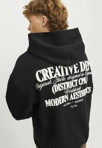 Zwarte hoodie met een grote witte print op de achterkant met verschillende tekstelementen. Het materiaal lijkt dik en textuur te hebben, met een ruime capuchon.