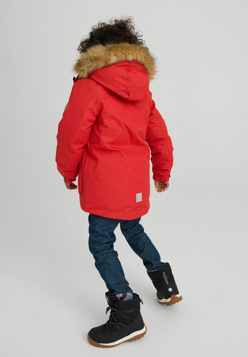 Chaqueta parka roja con capucha de piel sintética, mangas largas y bolsillos laterales. Combinada con vaqueros azul oscuro y botas de invierno negras.