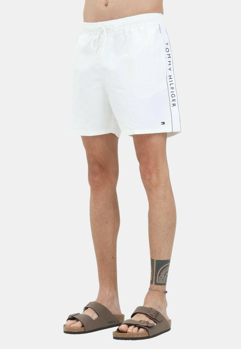Costume da bagno bianco con cintura elastica e accentuazione del logo sui lati in blu navy. Abbinato a sandali marroni con design a doppia fascia.