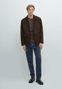 Blazer in suede marrone con chiusura a un bottone, abbinato a una t-shirt scura e jeans in denim blu. Il modello sta in piedi con le mani rilassate ai fianchi.