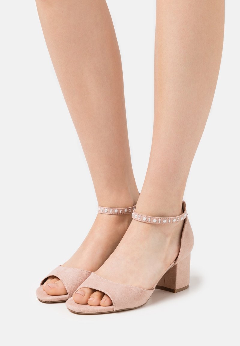 Pieds portant des sandales à talons carrés en suède rose clair avec des brides de cheville ornées de petits clous blancs, orteils partiellement visibles.