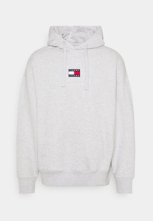 Felpa grigia chiara realizzata in misto cotone, con tasca frontale, cordini e un'etichetta logo rettangolare nei colori rosso, bianco e blu.