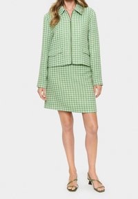 Zelená károvaná bunda se zapínáním na zip, k ní ladící sukně a sandály na podpatku. Látka má texturovaný vzor a strukturovaný střih.