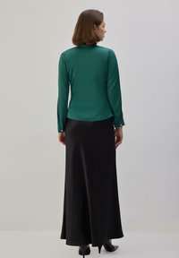 Mujer con cabello castaño hasta los hombros, vestida con una blusa de manga larga color verde azulado y una falda larga negra, de pie frente a una pared lisa y clara.