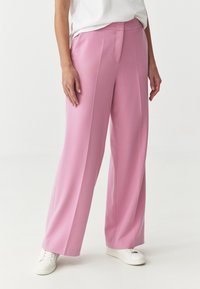 Roze, wijde broek van soepel stof met een subtiele glans. Voorzien van een plissé aan de voorkant en een platte tailleband. Gecombineerd met witte sneakers.
