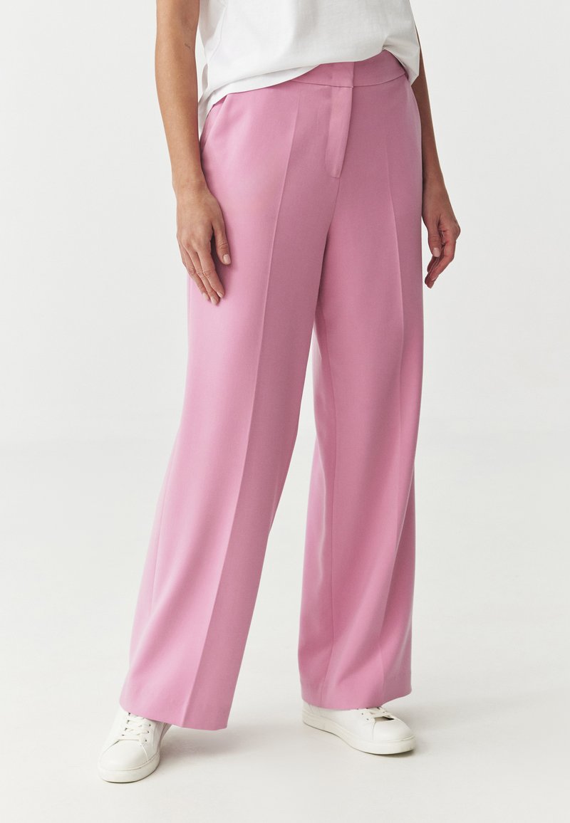 Roze, wijde broek van soepel stof met een subtiele glans. Voorzien van een plissé aan de voorkant en een platte tailleband. Gecombineerd met witte sneakers.