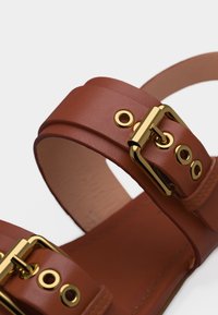 Sandale en cuir marron avec deux boucles en or et des œillets métalliques sur un fond blanc uni.