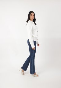 Cardigan blanco con canales y botones, combinado con jeans oscuros de tiro acampanado y tacones claros de punta abierta. Diseño simple y materiales texturizados.