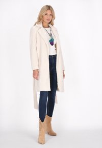 Cappotto lungo e peloso di colore crema con colletto, indossato sopra una maglietta bianca e jeans scuri. Presenta stivali cowboy color marrone chiaro con intagli decorativi.