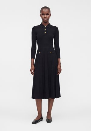 Donna che indossa un vestito midi nero plissettato a maniche lunghe con bottoni dorati sul petto e tasche, in piedi davanti a uno sfondo bianco semplice.