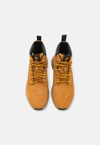 Bruna mockaskinnsankleboots med svarta detaljer, gula och röda snören, rund tå och gummisula, med en logotyp på tungan.