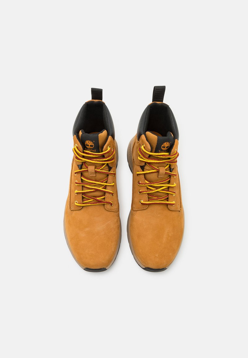 Botas de ante marrón hasta el tobillo con acentos negros, cordones amarillos y rojos, puntera redonda y suela de goma, con un logo en la lengüeta.