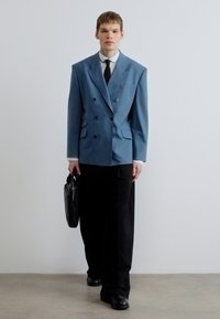 Sacou albastru cu dungi fine şi închidere dublă, croială supradimensionată, combinat cu pantaloni negri. Modelul ţine o servietă neagră şi poartă o cămaşă albă şi cravată.