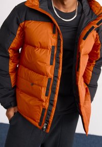Veste matelassée isolante en orange et noir, avec fermeture éclair, plusieurs panneaux cousus et poches extérieures. Texture douce.