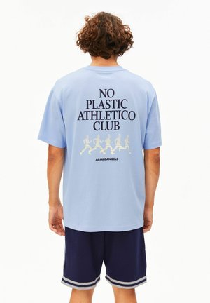 Persoon met bruin krullend haar die een lichtblauw T-shirt draagt met daarop de tekst "NO PLASTIC ATHLETICO CLUB" en hardlopersfiguren op de rug.
