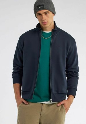 Felpa blu navy con zip e colletto alto, abbinata a una maglietta verde. Presenta una vestibilità slim e polsini a coste. Berretto grigio in testa.