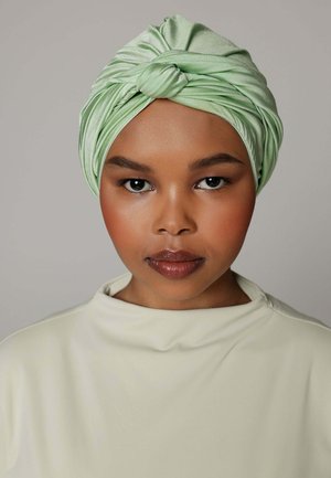 Jonge vrouw met een warme huidtint die een glanzende lichtgroene tulband en een crèmekleurige top met hoge hals draagt, kijkt recht vooruit tegen een effen achtergrond.