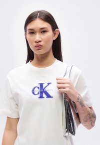 Λευκό βαμβακερό t-shirt με μπλε λογότυπο "CK" και κείμενο "Calvin Klein". Το μοντέλο κρατά μια μεταλλική ασημένια τσάντα. Ορατές λεπτομέρειες τατουάζ στο χέρι.