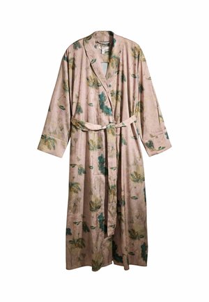 Frau steht barfuß vor einem sonnenbeschienenen Raum und trägt einen langen, floralen Bademantel, der in der Taille gebunden ist.