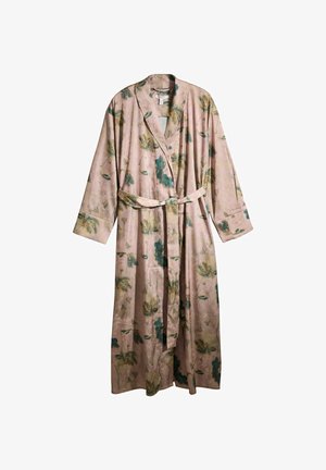 Femme debout pieds nus devant une pièce ensoleillée, portant une longue robe de chambre à motif floral nouée à la taille.