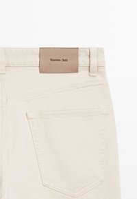 Beige Denim-Jeans mit Gesäßtasche, die eine gestickte Detailierung aufweist, sowie einem tanfarbenen Lederetikett mit der Aufschrift "Massimo Dutti" über der Tasche.