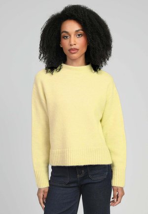 Femme aux cheveux bouclés portant un pull en maille jaune clair et un jean bleu foncé sur un fond clair uni.