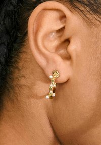 Guld hoops ørestikker har et tekstureret design med små hvide perler fastgjort. Ørestikkerne vises tæt på et øre.