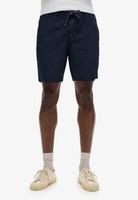 Shorts de bain navy avec un motif à pois blancs, taille élastique et cordon de serrage, complétés par des baskets et des chaussettes de couleur claire.