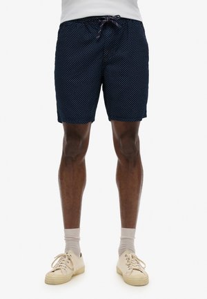 Marineblå svømmedragter med et hvidt prikket mønster, elastisk talje og snøre, suppleret med lyse sneakers og sokker.