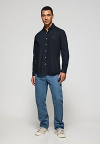 Camisa de mezclilla azul oscuro con mangas largas, tejido de algodón, cuello clásico y pequeño logotipo rojo en el pecho; combinada con jeans azules y zapatillas deportivas.
