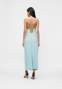 Γυναίκα με μακριά σκουρόχρωμα μαλλιά, φορώντας ανοιχτό γαλάζιο φόρεμα χωρίς πλάτη με λεπτούς διασταυρούμενους ιμάντες και μαύρα ψηλοτάκουνα σανδάλια, στέκεται σε μονόχρωμο φόντο.