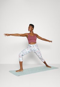 Top cropped em bordô e leggings estampadas com flores em cores pastel. Pessoa a realizar uma pose de ioga sobre um tapete azul claro.