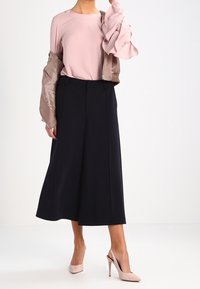 Blusa rosa chiaro con maniche arricciate, abbinata a culotte blu navy a gamba larga e scarpe con tacco beige. Texture morbide e design moderno.