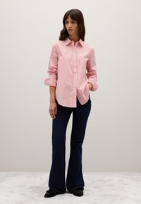 Una donna è in piedi indossando una camicia a righe rosa e bianca con le maniche arrotolate, jeans a zampa d'elefante blu scuro e scarpe nere.