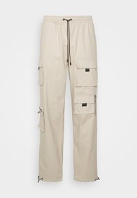 Beige cargo kalhoty s několika kapsami, stahovací pas, elastické manžety a zipový detail na boku. Vyrobeno z odolného materiálu.