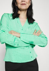 Blusa verde, semi-transparente, com decote em V, fecho com botões, mangas longas e barra estilo peplum; apresenta padrões de pontos refletivos por toda a peça.
