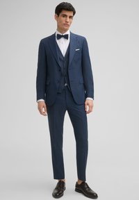 Marineblauer Dreiteiler aus strukturiertem Stoff, mit einem Spitzrevers, einer Weste und einem weißen Hemd, ergänzt durch eine schwarze Fliege und braune Schuhe.