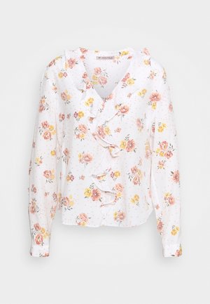 Witte bloemenblouse met roze en gele bloemen, ruches langs de halslijn en lange mouwen. Zacht materiaal met een lichte textuur.