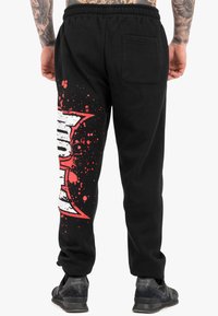 Schwarze Jogginghose mit einem rot-weißen Grafikdruck, der Spritzmuster zeigt. Elastischer Bund und Gesäßtasche für funktionale Verwendung.