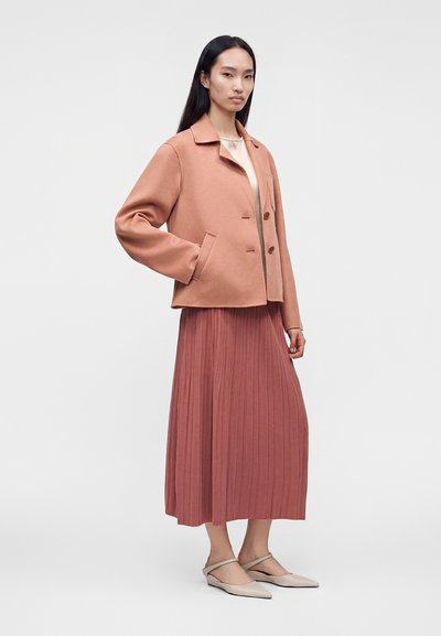 Femme portant une veste marron clair boutonnée par-dessus un haut crème et une longue jupe plissée couleur rouille avec des chaussures beige pointues.