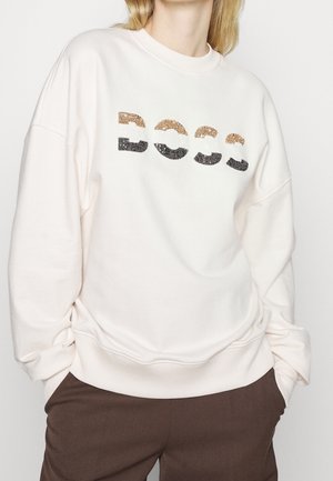 Person iført en hvid sweatshirt med palietter i abstrakte bogstaver og brune bukser, hænderne i lommerne, mod en ensfarvet baggrund.