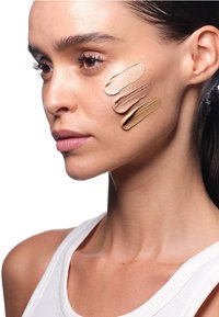 Quattro tonalità di fondotinta in diverse sfumature di beige chiaro sono applicate in una linea sulla guancia del modello, mostrando variazioni di texture e colore.